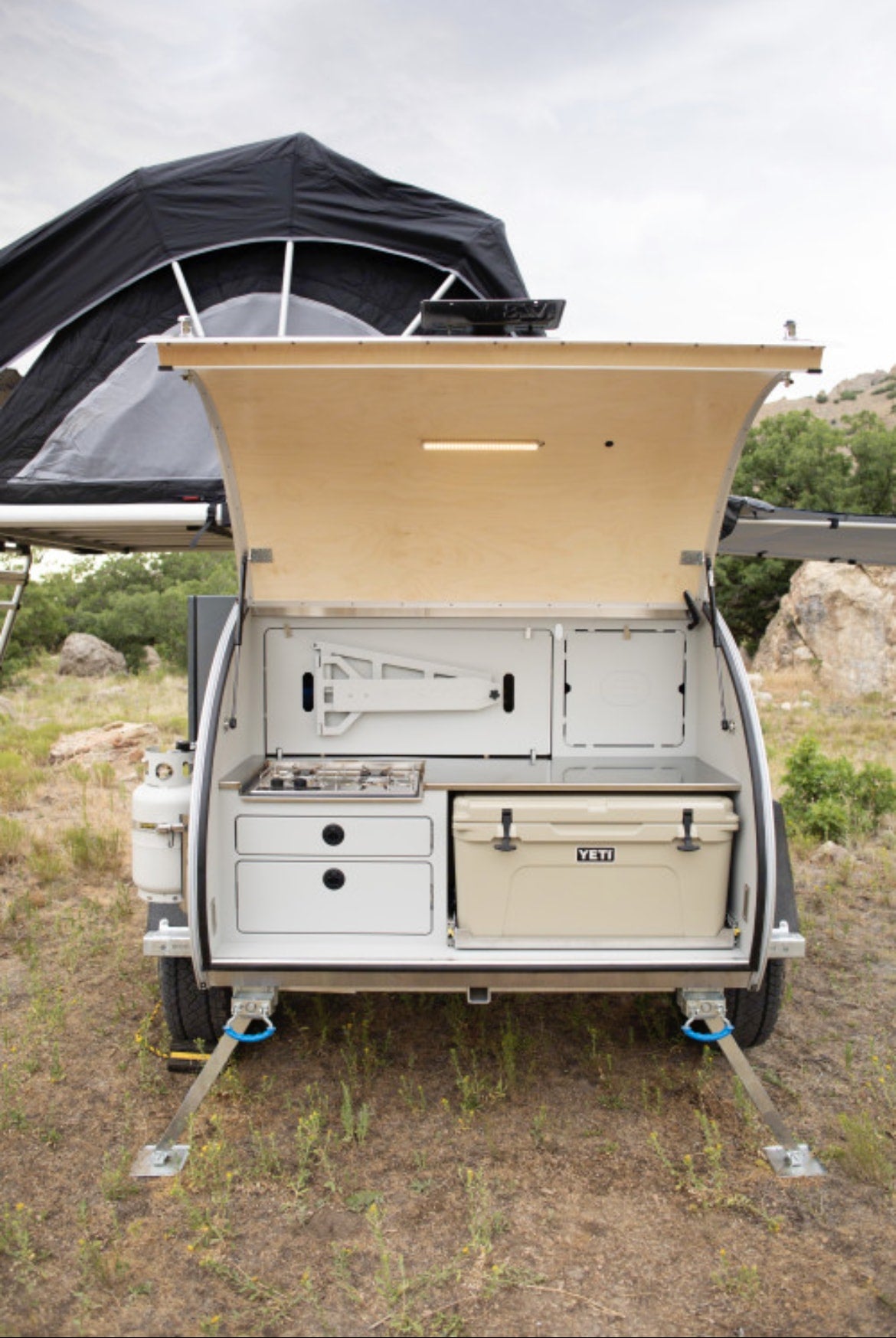  featuring an awning and off-road tires.|Experience outdoor adventure with an RV rental: a teardrop camper parked in a desert landscape with a dog and campfire.|Compact RV rental teardrop trailer shows its rear outdoor kitchen: stove — photo 22