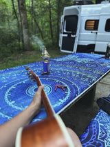 Relaxing at an RV rental campsite: playing guitar & enjoying calming incense on a vibrant blue table, with a camper van in the forest.