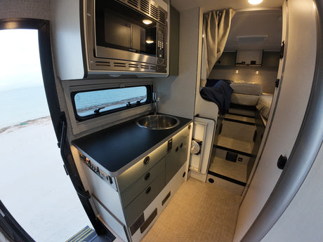 This RV rental interior features a compact kitchen with a microwave, sink, and a cozy bed area, overlooking a sandy beach.