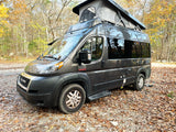 A grey Ram ProMaster camper van with a pop-top roof is parked on a leaf-strewn path in an autumn forest, ready for rv rental.