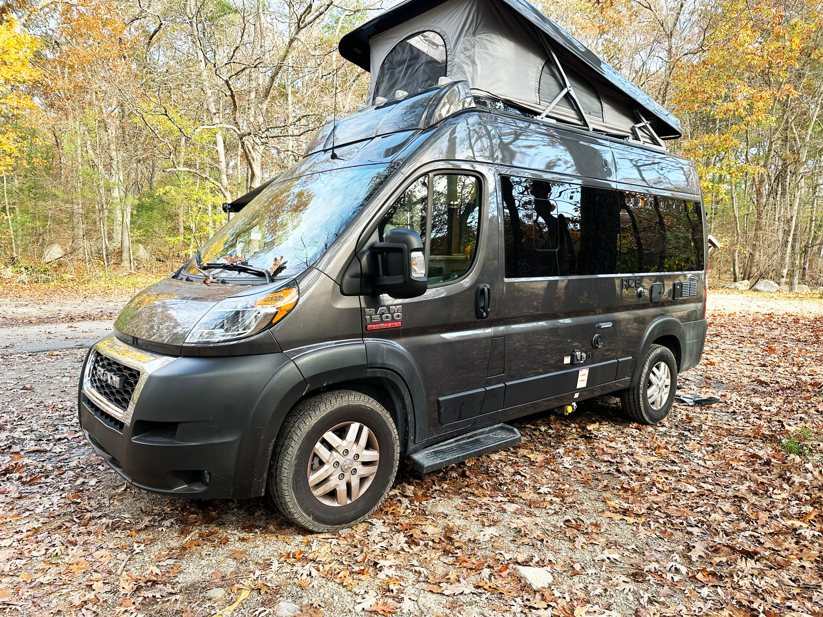 A grey Ram ProMaster camper van with a pop-top roof is parked on a leaf-strewn path in an autumn forest, ready for rv rental.