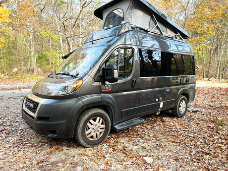 A grey Ram ProMaster camper van with a pop-top roof is parked on a leaf-strewn path in an autumn forest, ready for rv rental.