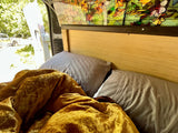 Cozy RV rental bed with gold blanket, gray pillows, stained glass & open door view.