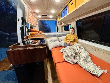 Cozy camper van interior for your next rv rental adventure. Woman smiles on bench with kitchen & bed.