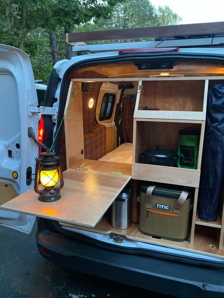Cozy camper van interior: plaid walls, custom shelves, pull-out table with lantern, and camping gear. Ideal for rv rental adventures.
