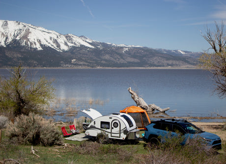 An RV rental teardrop camper and SUV are set up for camping by a tranquil lake, backed by snow-capped mountains under clear skies.