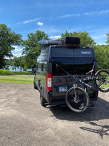 Winnebago rv rental: Dark grey camper van with a bike rack parked near a scenic lake on a sunny day.