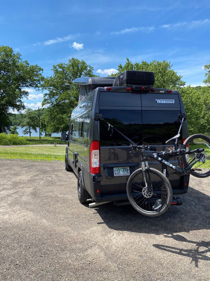 Winnebago rv rental: Dark grey camper van with a bike rack parked near a scenic lake on a sunny day.