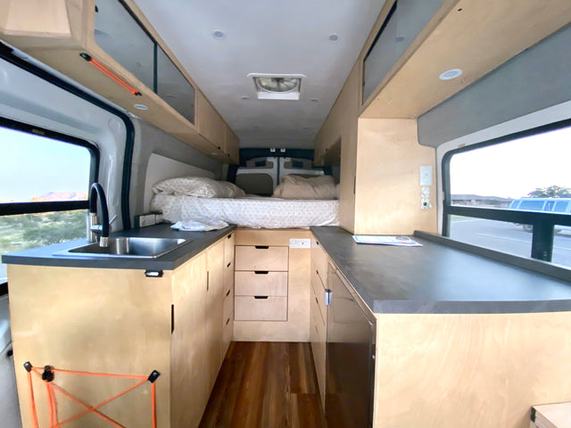 Modern camper van RV rental interior, featuring a compact kitchen, bed, and custom wood cabinetry.