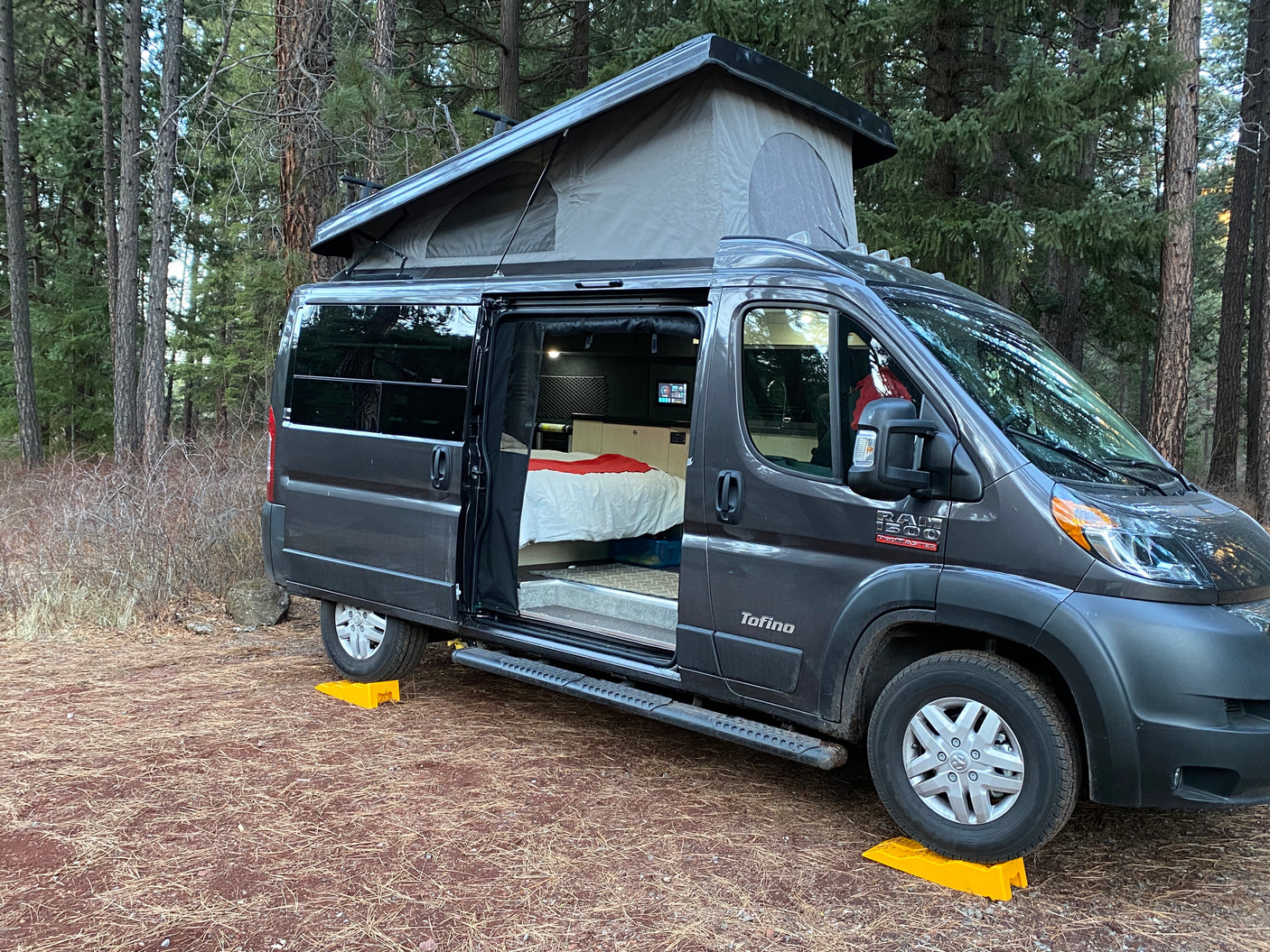 An rv rental, a grey camper van with a pop-top, parked in a forest clearing, side door open showing the bed. Ready for adventure!