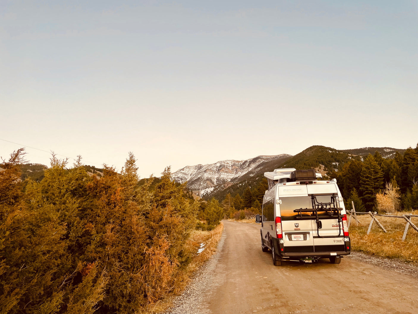 An RV rental camper van rests on a dirt road, flanked by evergreen trees and snow-dusted mountains under a clear sky.