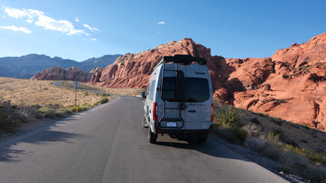 A grey Winnebago RV rental on a desert road, flanked by vibrant red rock mountains under a clear blue sky.