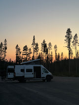 White RV rental camper van with bikes, parked in a forest at sunset with a vibrant orange and blue sky.
