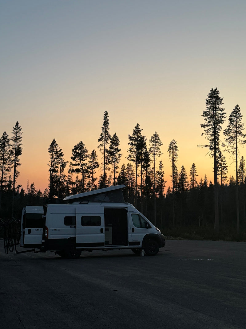 White RV rental camper van with bikes, parked in a forest at sunset with a vibrant orange and blue sky. — photo 2