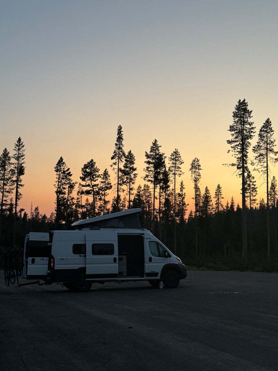 White RV rental camper van with bikes, parked in a forest at sunset with a vibrant orange and blue sky.
