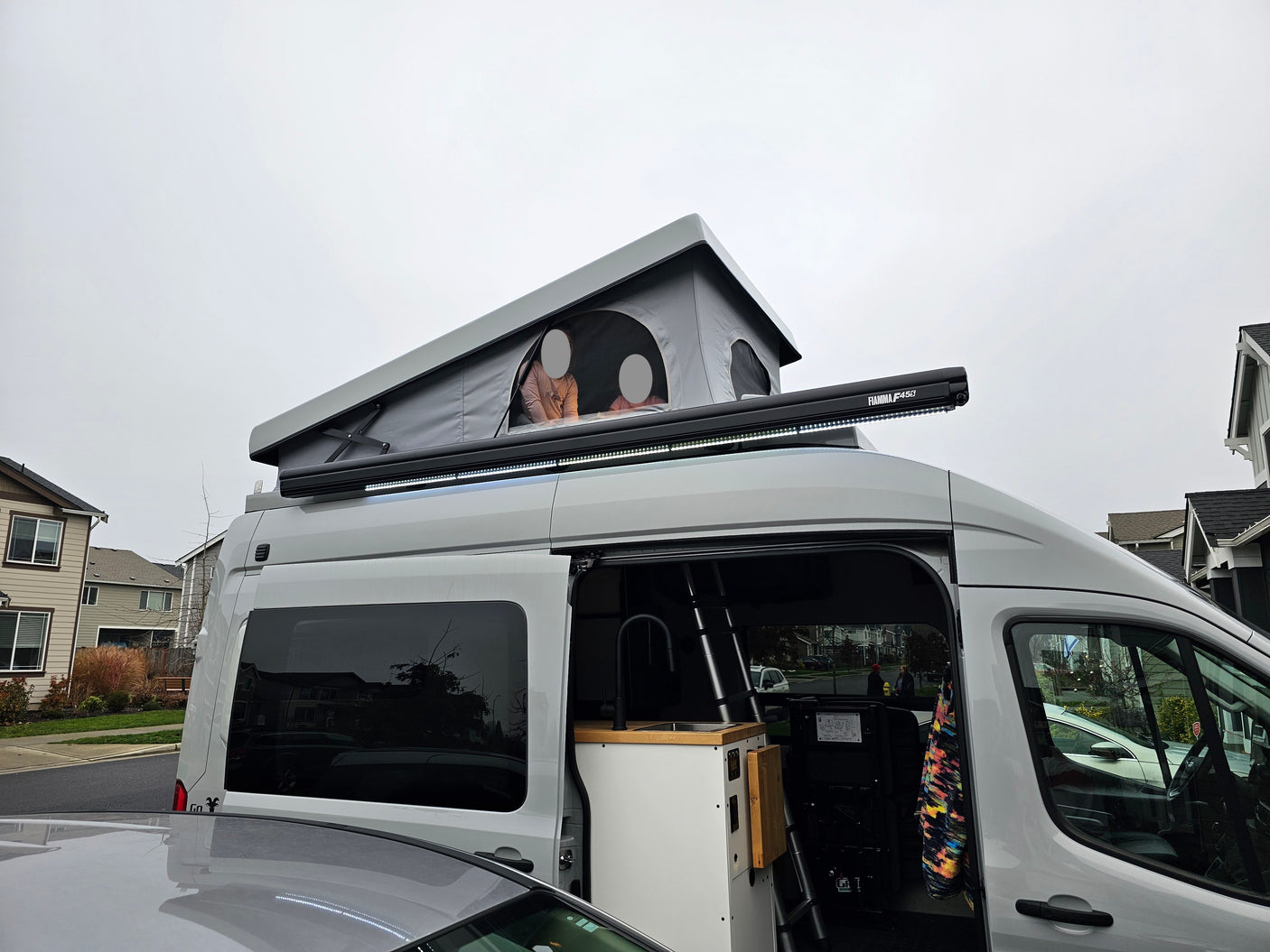 Gray rv rental with open pop-top tent (people inside), exterior awning, and compact interior kitchenette. Ideal for adventure.