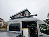 Gray rv rental with open pop-top tent (people inside), exterior awning, and compact interior kitchenette. Ideal for adventure.