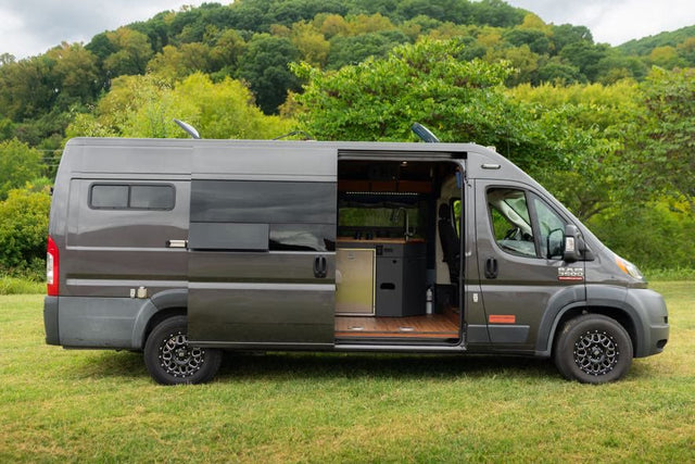 Gray Ram ProMaster camper van, ideal for RV rental adventures, parked on grass with its side door open showing the interior kitchen.