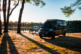Black camper van (an RV rental) by a serene lake at sunset, with long shadows from nearby trees. Perfect for outdoor adventures.
