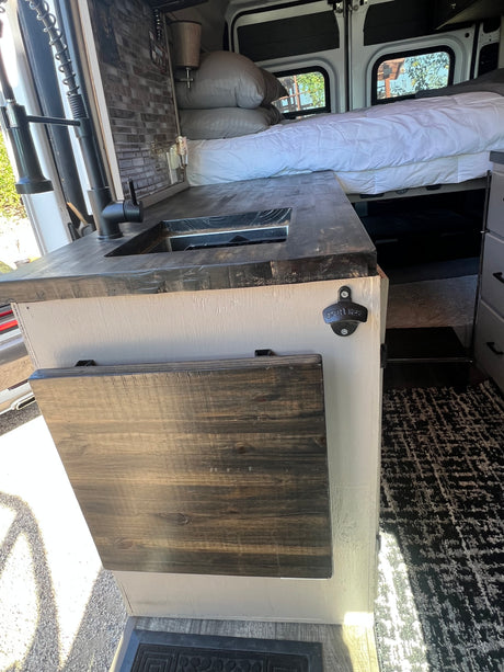 Inside this RV rental: kitchenette with sink, fold-down table, bottle opener, and a cozy bed with white bedding.