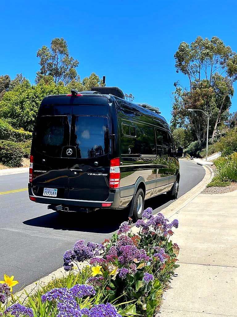 Black Mercedes-Benz Sprinter RV, an ideal rv rental for travel, parked on a scenic road with vibrant flowers.