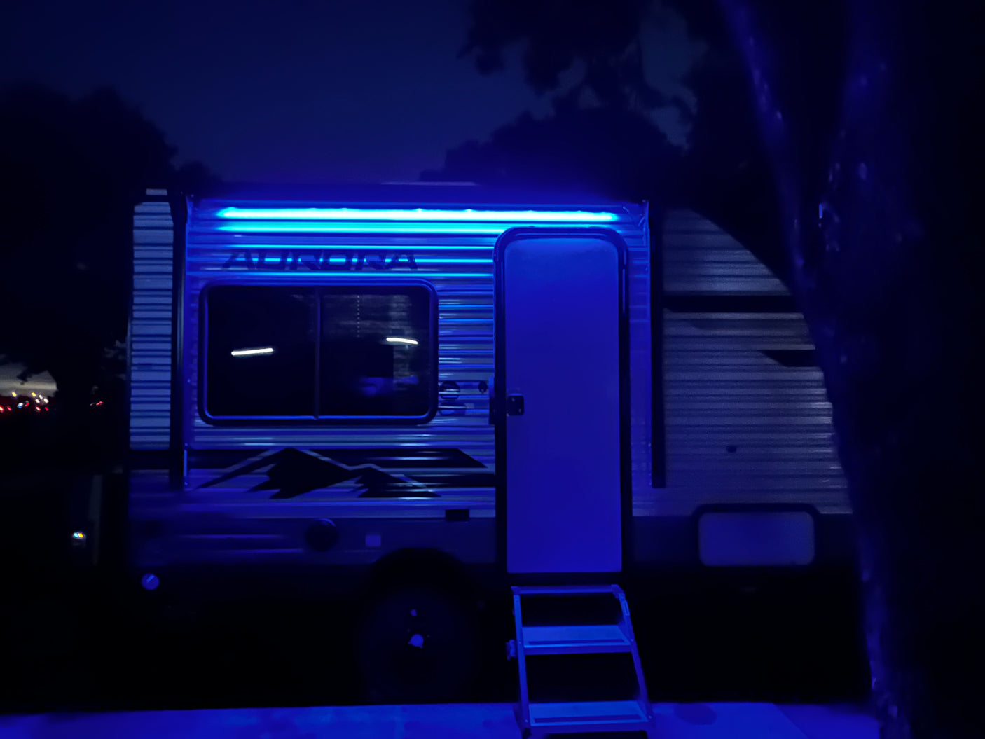 Bright blue LED lights illuminate this RV rental at night. The travel trailer's door is open, with steps leading inside for camping fun.