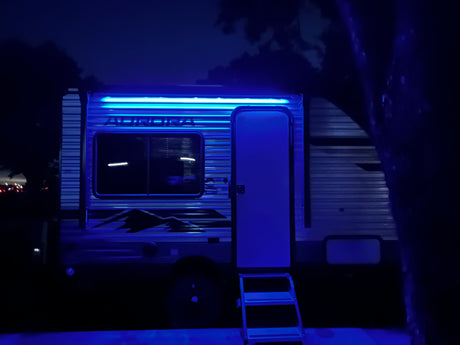 Bright blue LED lights illuminate this RV rental at night. The travel trailer's door is open, with steps leading inside for camping fun.