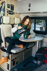 Woman pours coffee in a modern RV rental dinette. Control panels on wall, window view, perfect for mobile travel.