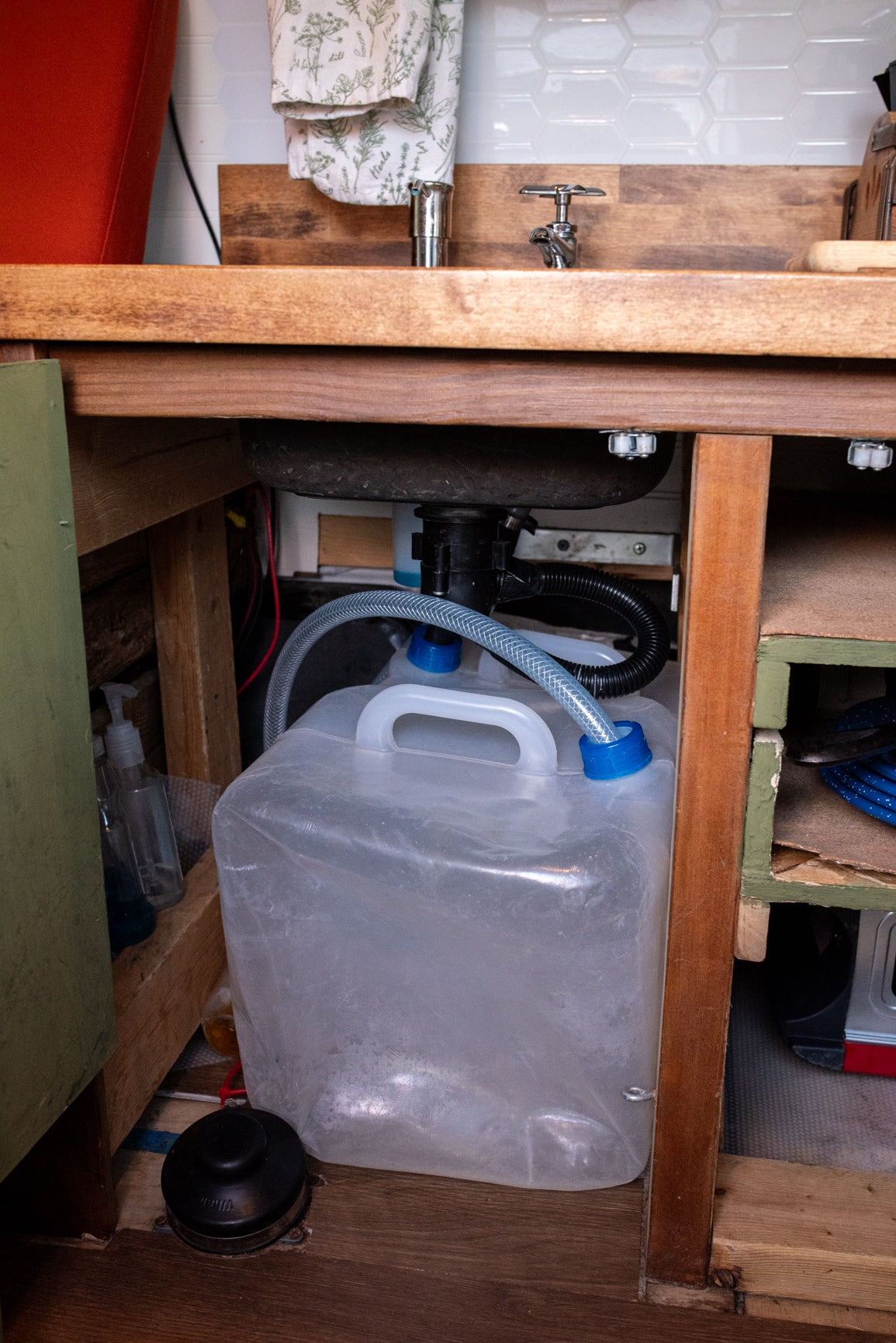 RV rental kitchen setup: under-sink view reveals a grey water collection jug and a floor pump for the fresh water faucet.