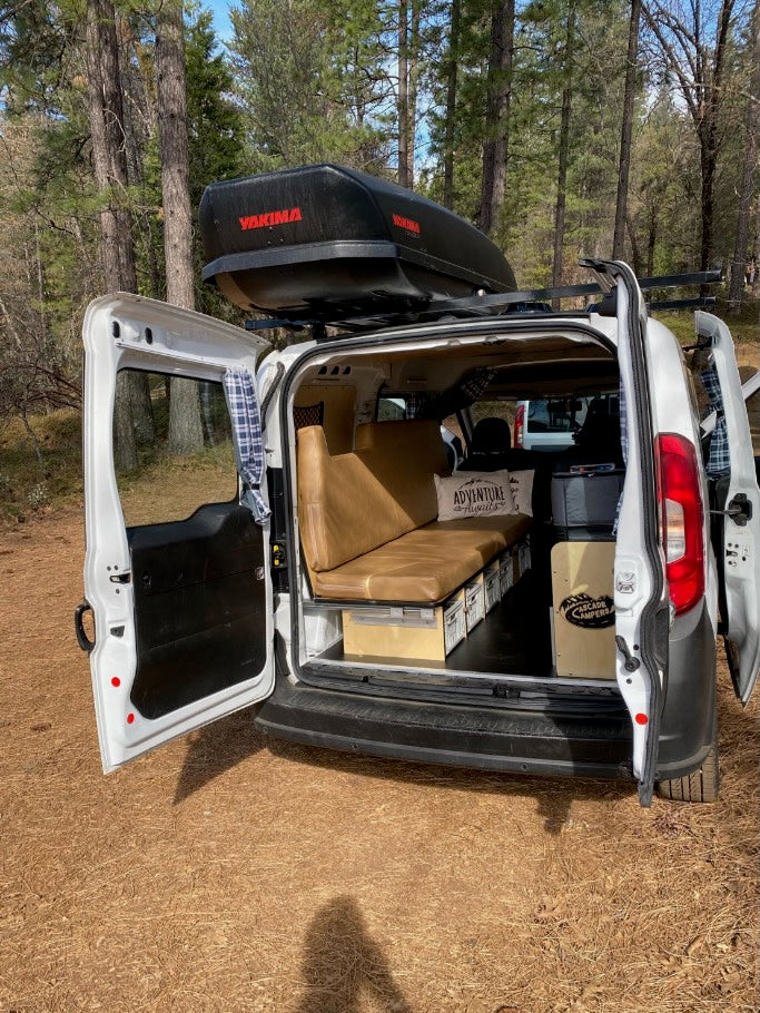 White camper van RV rental, backed into a forest clearing with its doors open, revealing a custom adventure interior and roof cargo box.