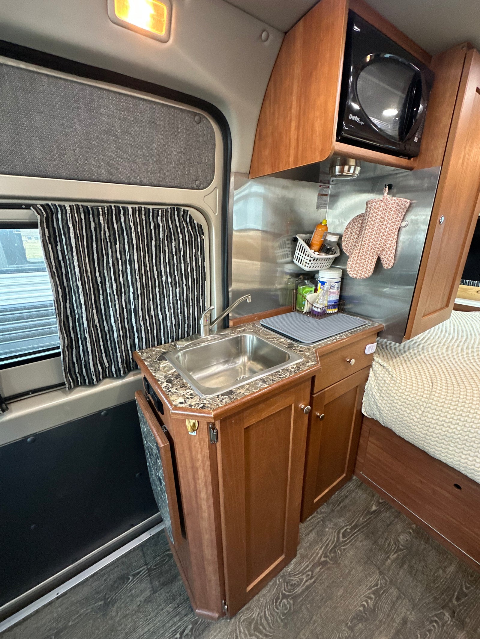 RV rental interior: compact kitchen with stainless sink, microwave, wood cabinets, striped curtains, and a comfy bed.