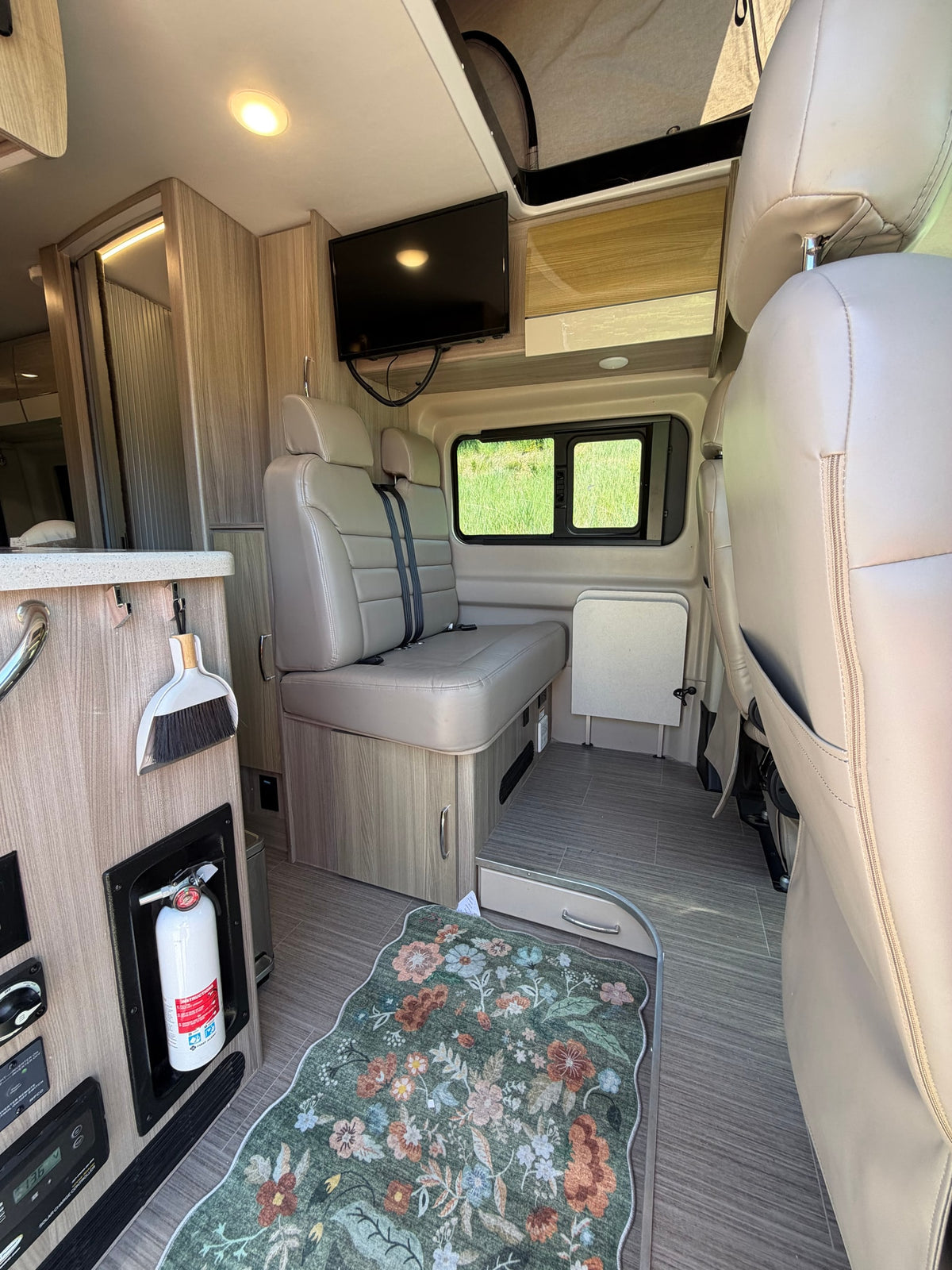 Cozy rv rental interior featuring tan seating, TV, built-in fire extinguisher, and a floral rug, with a view of green outside. — photo 6