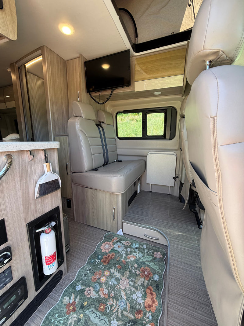 Cozy rv rental interior featuring tan seating, TV, built-in fire extinguisher, and a floral rug, with a view of green outside. — photo 6