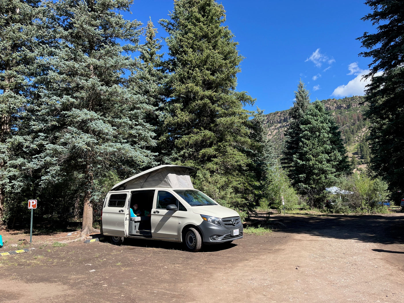 RV rental camper van with pop-top open at a rustic campsite. Surrounded by tall evergreens & mountains under a clear blue sky.