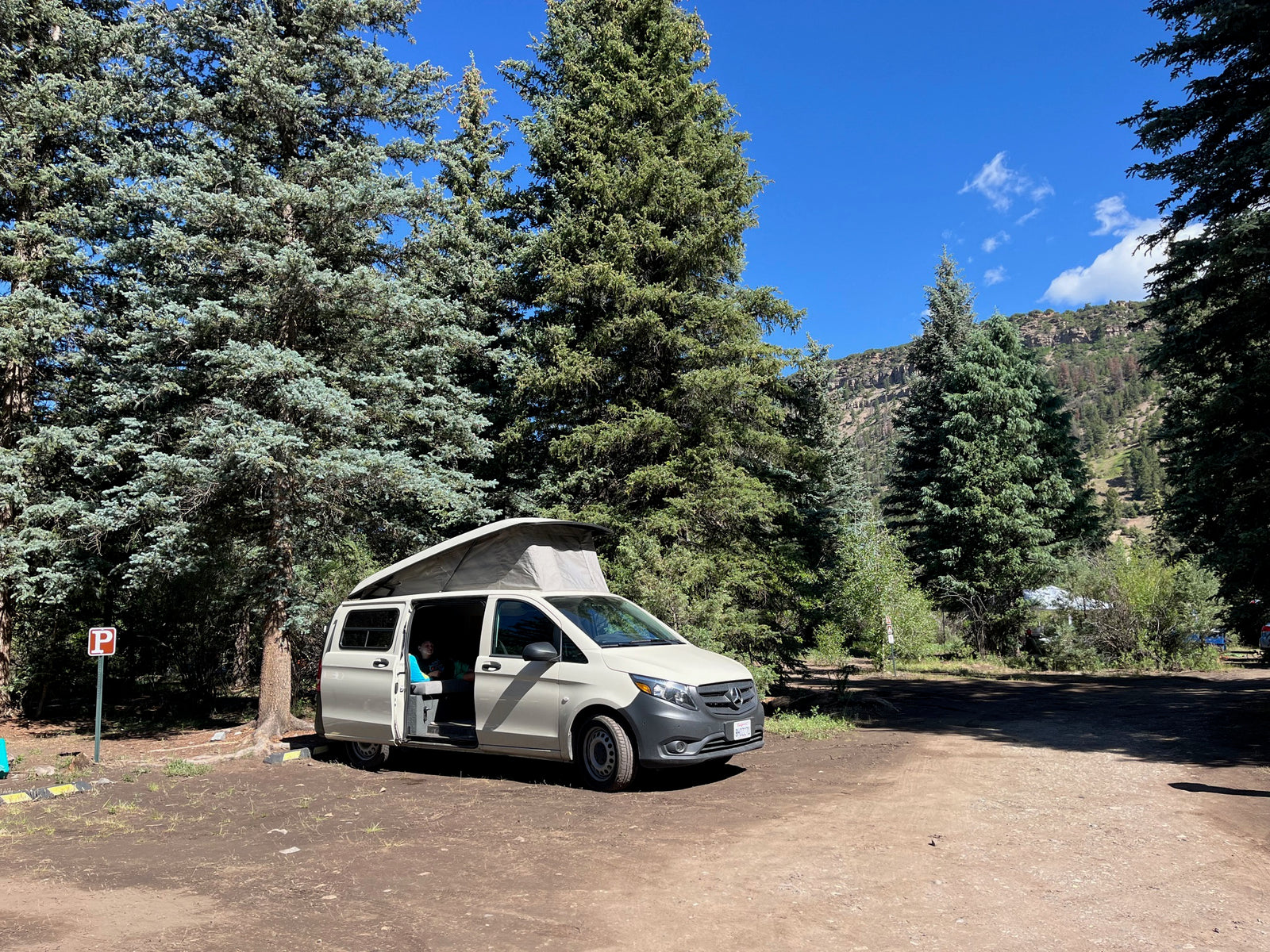 RV rental camper van with pop-top open at a rustic campsite. Surrounded by tall evergreens & mountains under a clear blue sky.