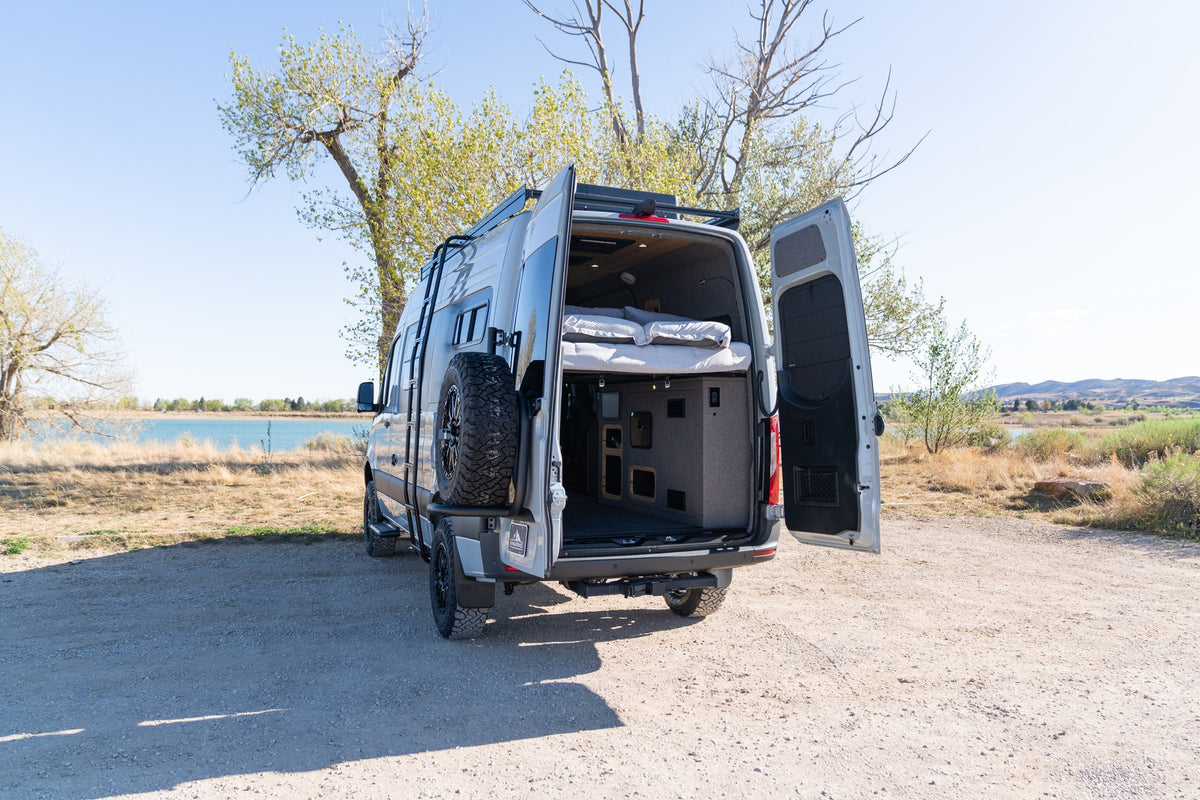An rv rental camper van with open rear doors reveals a cozy bed and storage, parked on a dirt path by a scenic lake. — photo 5