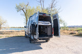 An rv rental camper van with open rear doors reveals a cozy bed and storage, parked on a dirt path by a scenic lake.