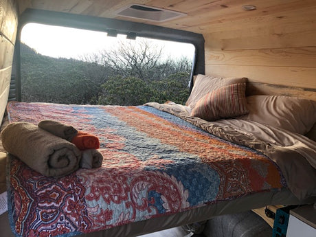 Comfortable rv rental bed with colorful quilt, rolled towels, and a beautiful natural view from the large window.