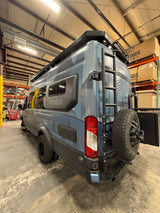 Rear view of a custom gray & blue Vandoit camper van, with a ladder, spare tire, & robust bumper, for your next RV rental.