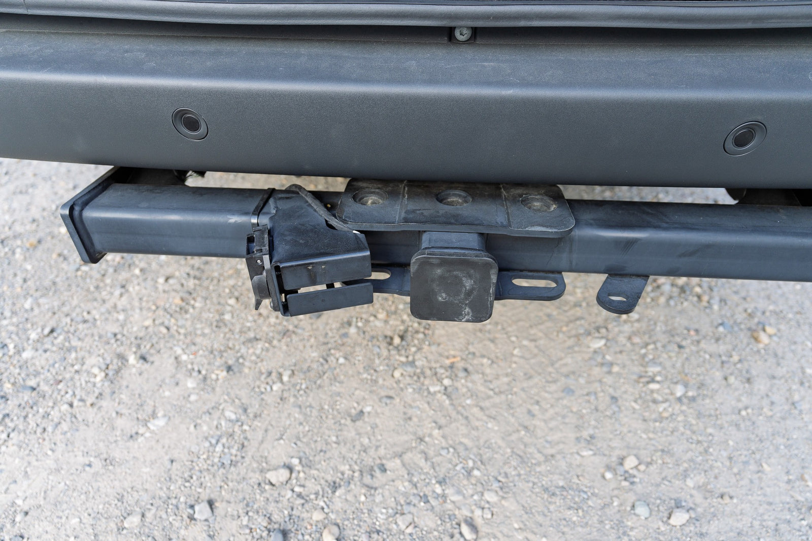 Close-up of an RV rental vehicle's rear tow hitch receiver with wiring harness and bumper parking sensors over gravel.