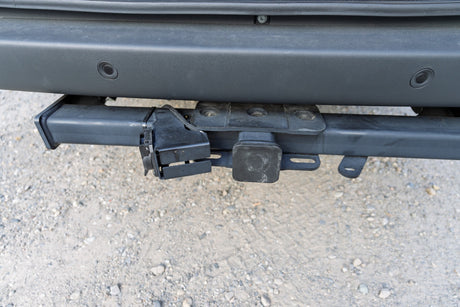 Close-up of an RV rental vehicle's rear tow hitch receiver with wiring harness and bumper parking sensors over gravel.