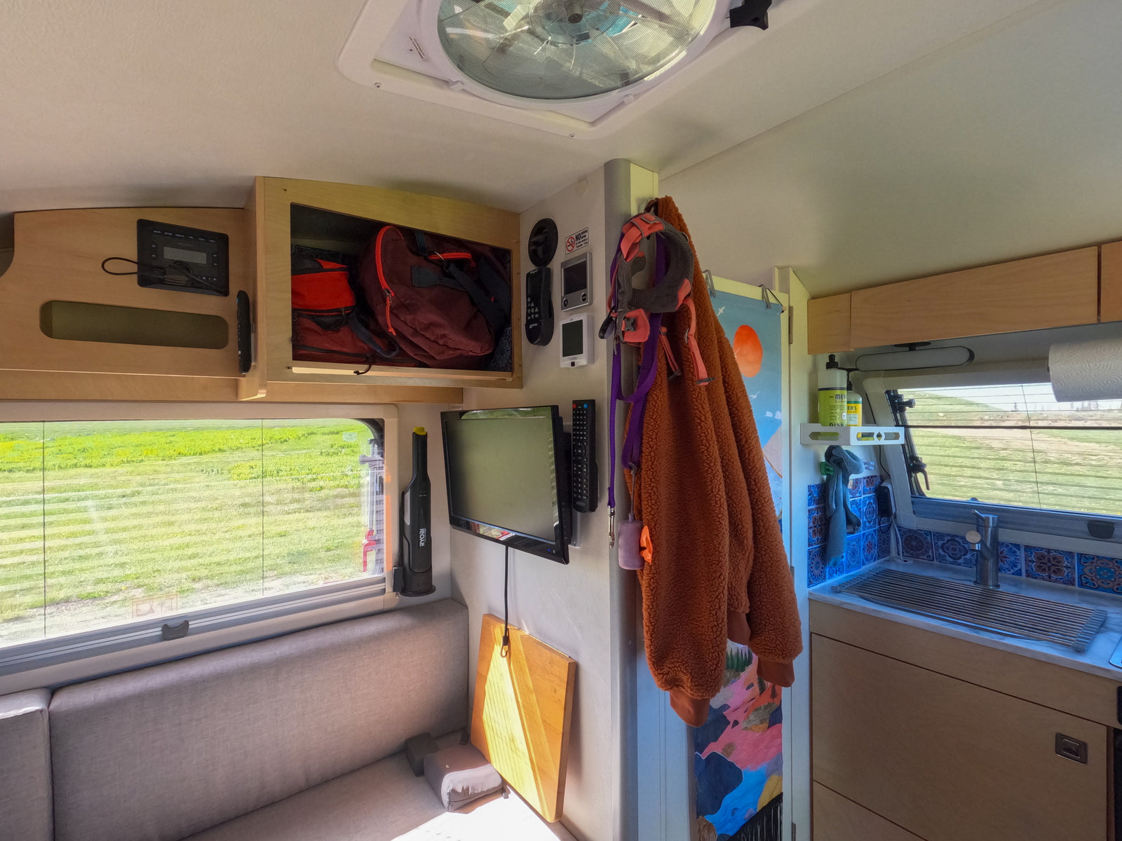 Bright RV rental interior, featuring a sofa, window with a view, TV, overhead storage, and a tiled kitchenette.