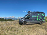 An Adventure Rig rv rental van, with green graphics & roof rack, parked on dry grass before the Grand Teton mountains.