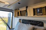 Inside a cozy RV rental: bed, overhead shelves, power outlets, and a screened window revealing outdoor nature.