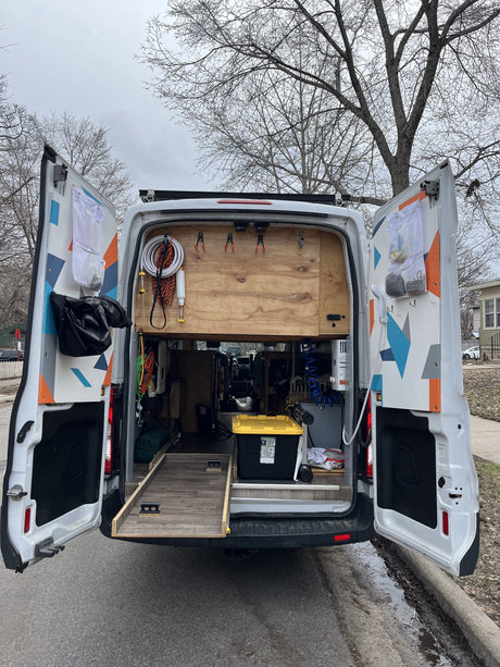 Open rear doors of an RV rental van reveal a custom-built interior with organized gear, an outdoor shower, and pull-out storage.