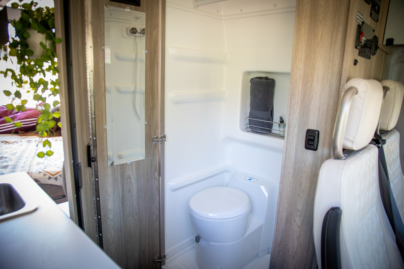 Compact RV rental interior: a white toilet, shower, and mirror in a small bathroom. Also shows sink, seats, and a bed.