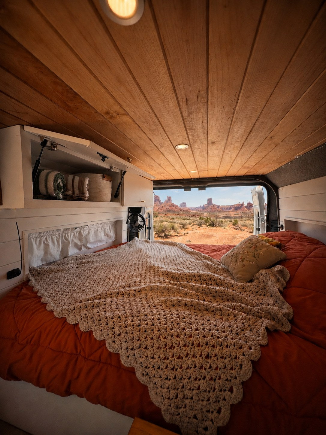 Inside an RV rental: a cozy bed, crochet blanket, wood ceiling, and breathtaking desert views of iconic rock formations.