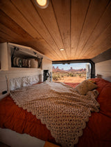 Inside an RV rental: a cozy bed, crochet blanket, wood ceiling, and breathtaking desert views of iconic rock formations.