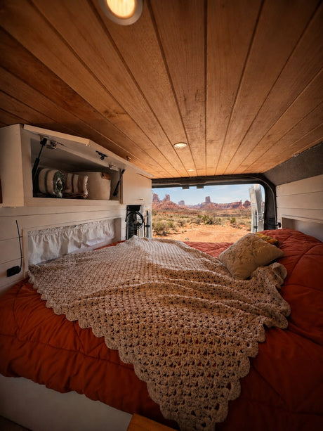 Inside an RV rental: a cozy bed, crochet blanket, wood ceiling, and breathtaking desert views of iconic rock formations.