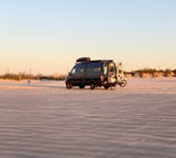 Enjoy an **rv rental** on the beach! Dark camper van with open doors, bike, & roof storage on wavy sand at golden hour.